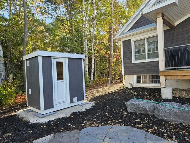 Grey sauna with white trim on a gravel pad beside a house