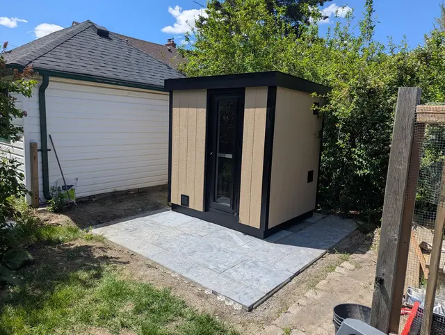 The Perhe Plus in tan and black, on a paver pad beside a garage