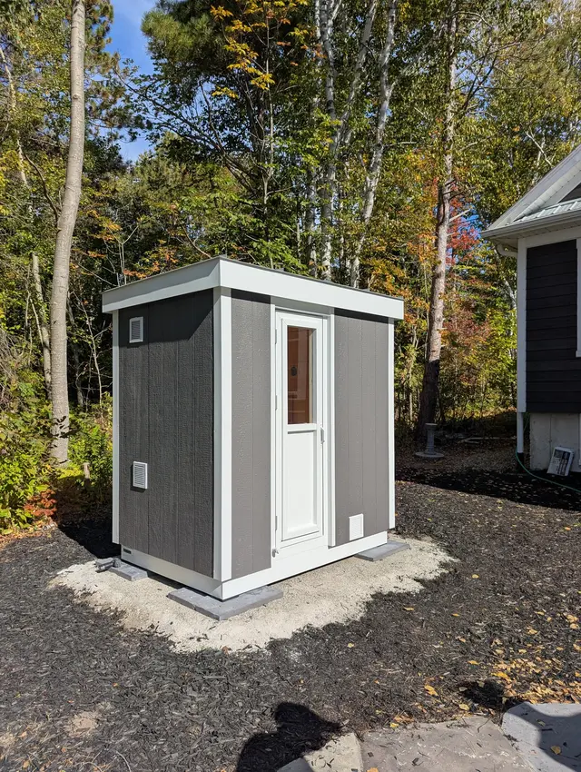 Grey sauna with white trim and a half glass door on a gravel pad