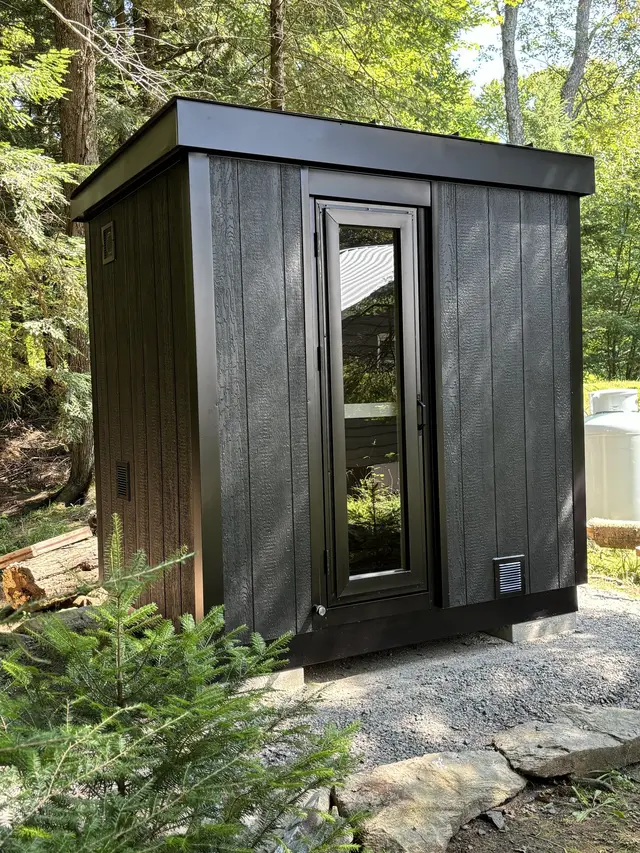 Black sauna with a glass door on a gravel pad beside rocks