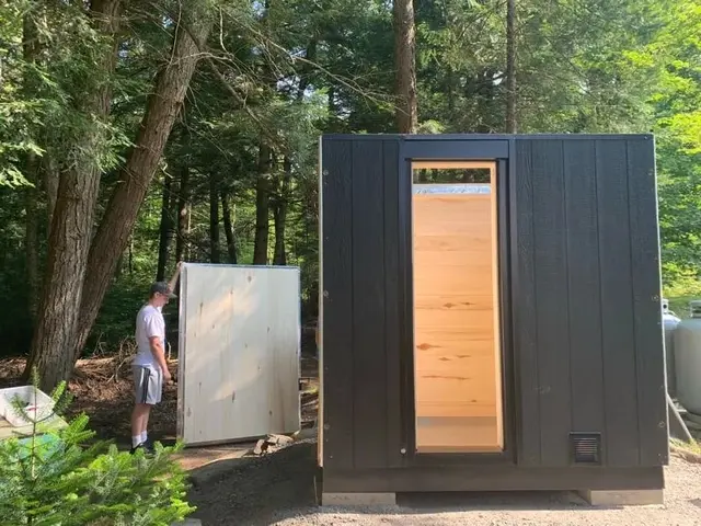 Black sauna with a glass door among trees, beside a stacked wall panel