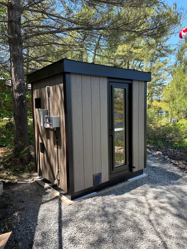 Taupe sauna with black trim and a glass door on a gravel pad
