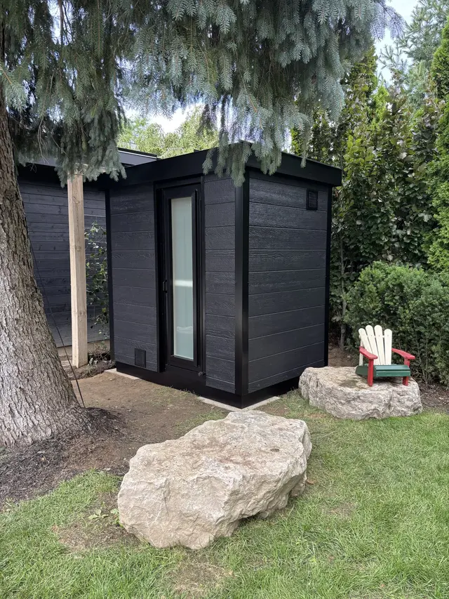 Black sauna in a garden beside a large boulder