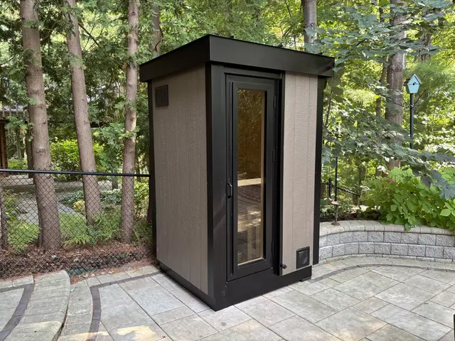 Taupe sauna with black trim and a glass door on a paver patio