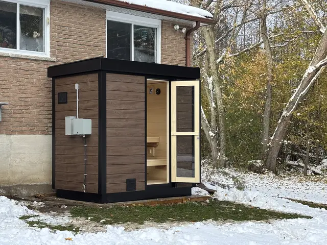 Woodgrain sauna with the door open, beside a house in the snow