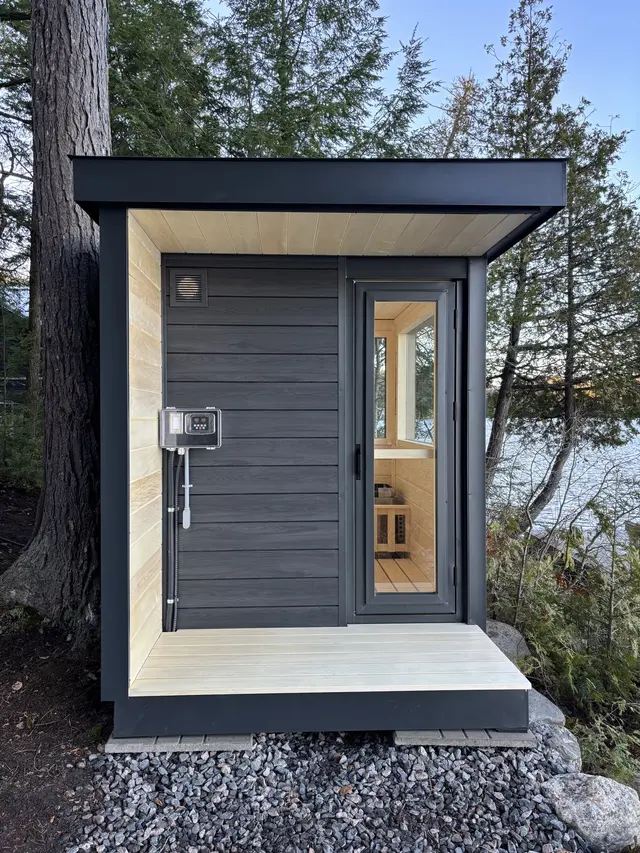 Grey sauna with a light wood porch and glass door on gravel by a lake