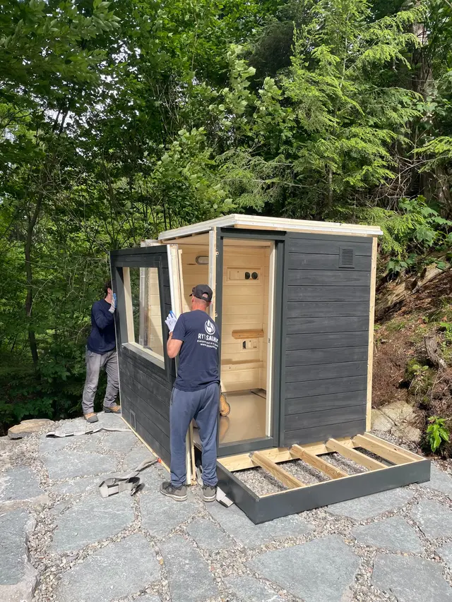 Installers assembling a sauna with a front window on a stone patio