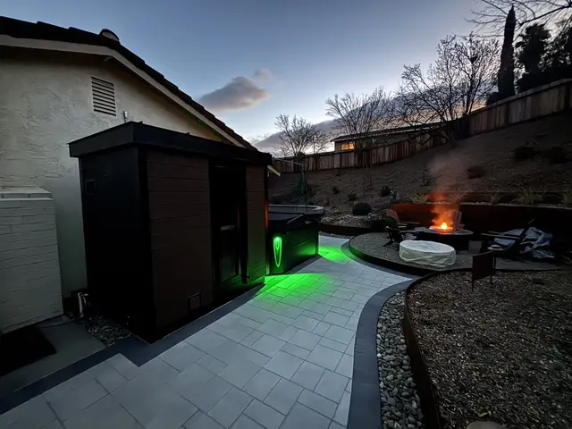 Black sauna with green lighting beside a patio and fire pit at dusk