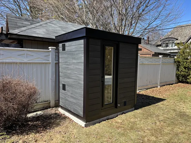 Grey and black sauna on a lawn beside a white fence