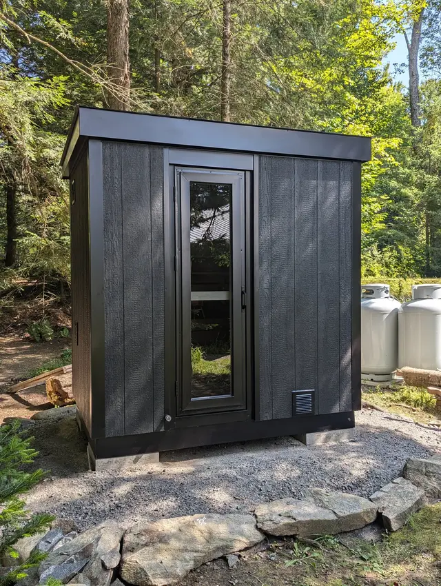 Black sauna with a glass door on a gravel pad in the woods