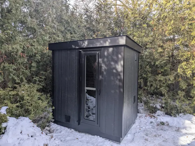 Black sauna in the snow in front of cedar hedges