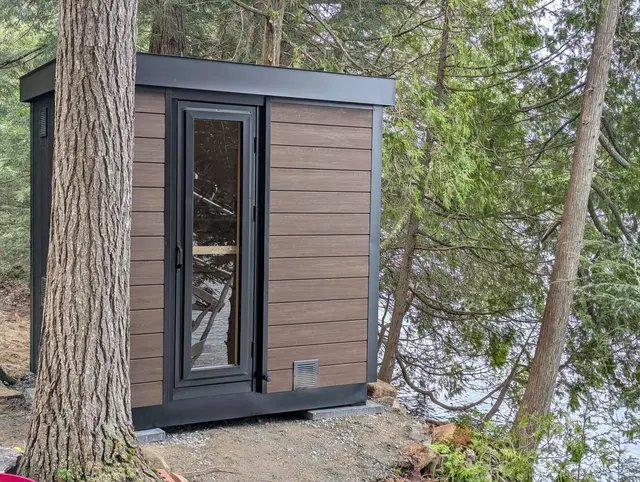 Brown sauna with black trim and a glass door beside a tree