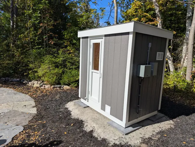 Grey sauna with white trim and an electrical box on a gravel pad