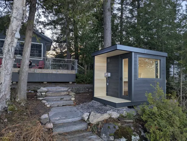 Grey sauna with a light wood porch and a front window, beside stone steps