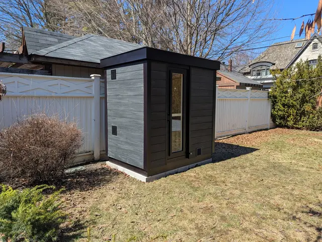 Grey and black sauna on a lawn beside a white fence