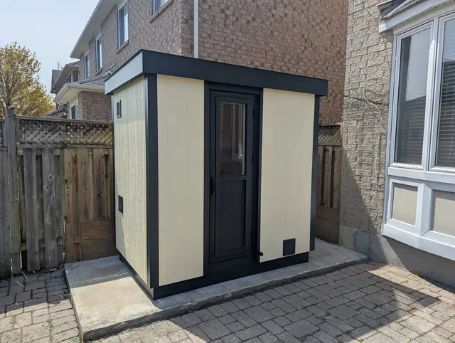 Cream and black sauna on a concrete pad beside a brick house
