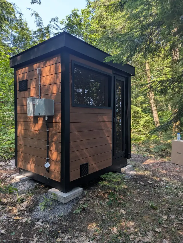 Woodgrain sauna with black trim, a side window and an electrical box, in a wooded yard