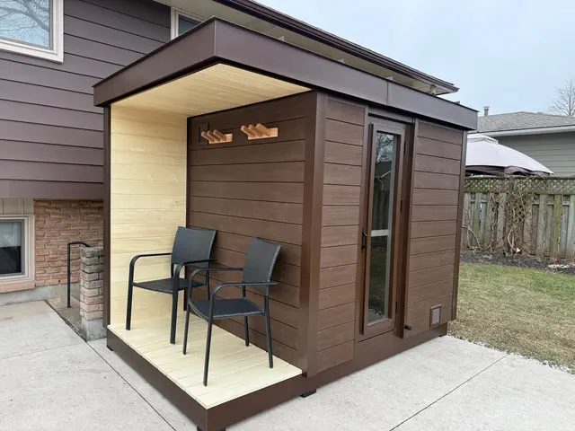 Brown sauna with a covered porch and two chairs, beside a house