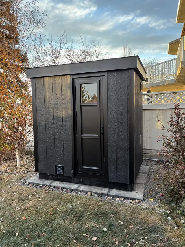 Black sauna with a small top glass door and an aluminum exterior, on a stone pad