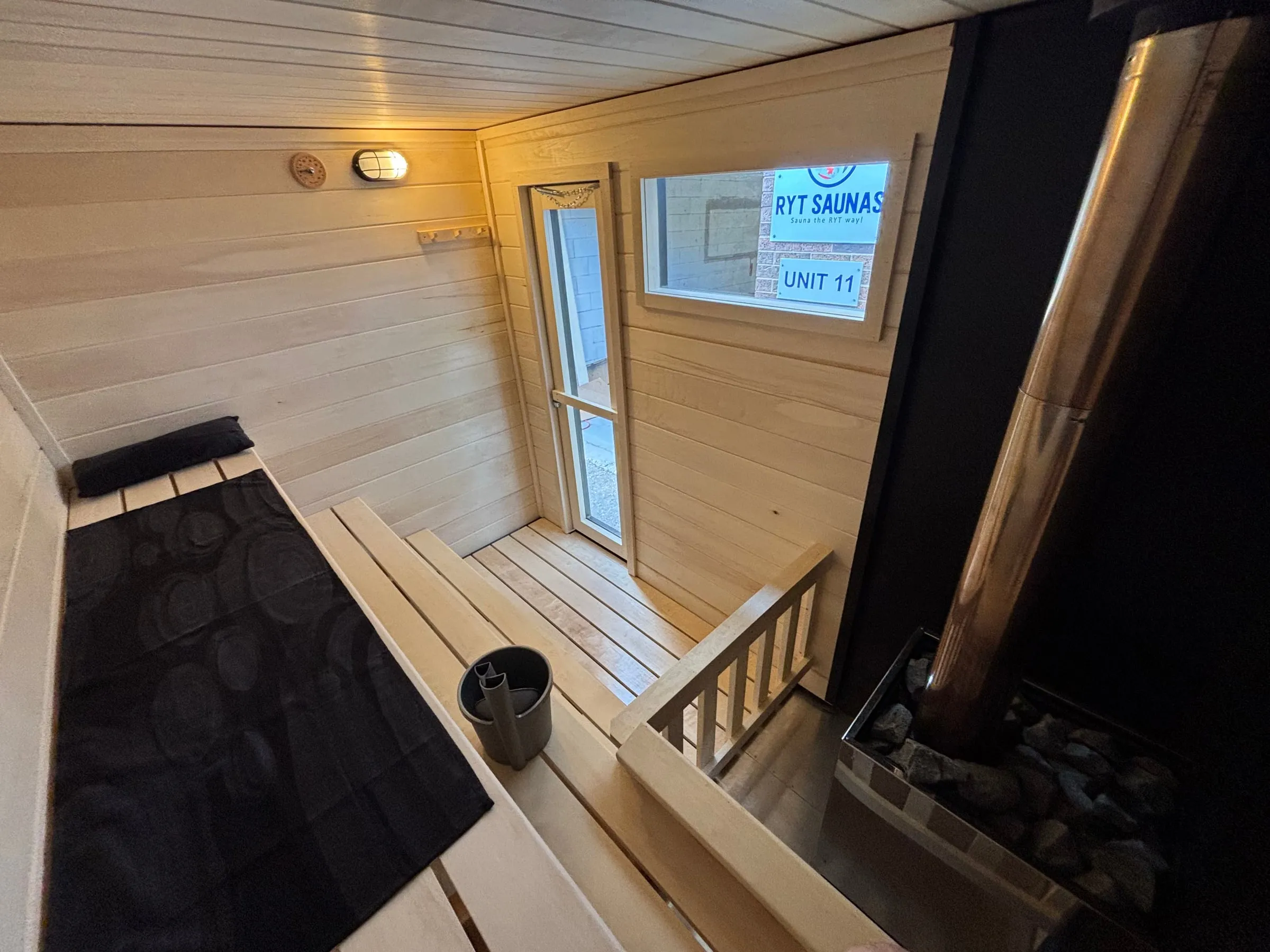 Inside our wood-burning trailer sauna: aspen benches with a towel and sauna bucket, the glass door and window, the chimney pipe and the heater rocks behind the guard rail.