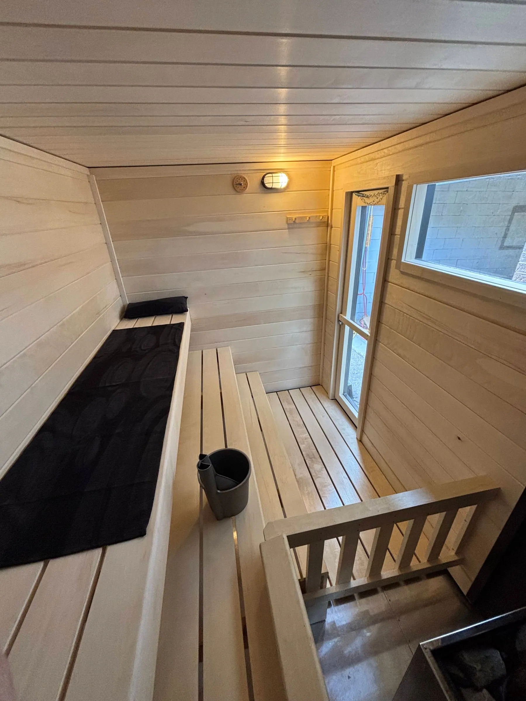 Inside our wood-burning trailer sauna: aspen benches with a towel and sauna bucket, a glass door and a window, a wall light, and the aspen guard rail around the heater.