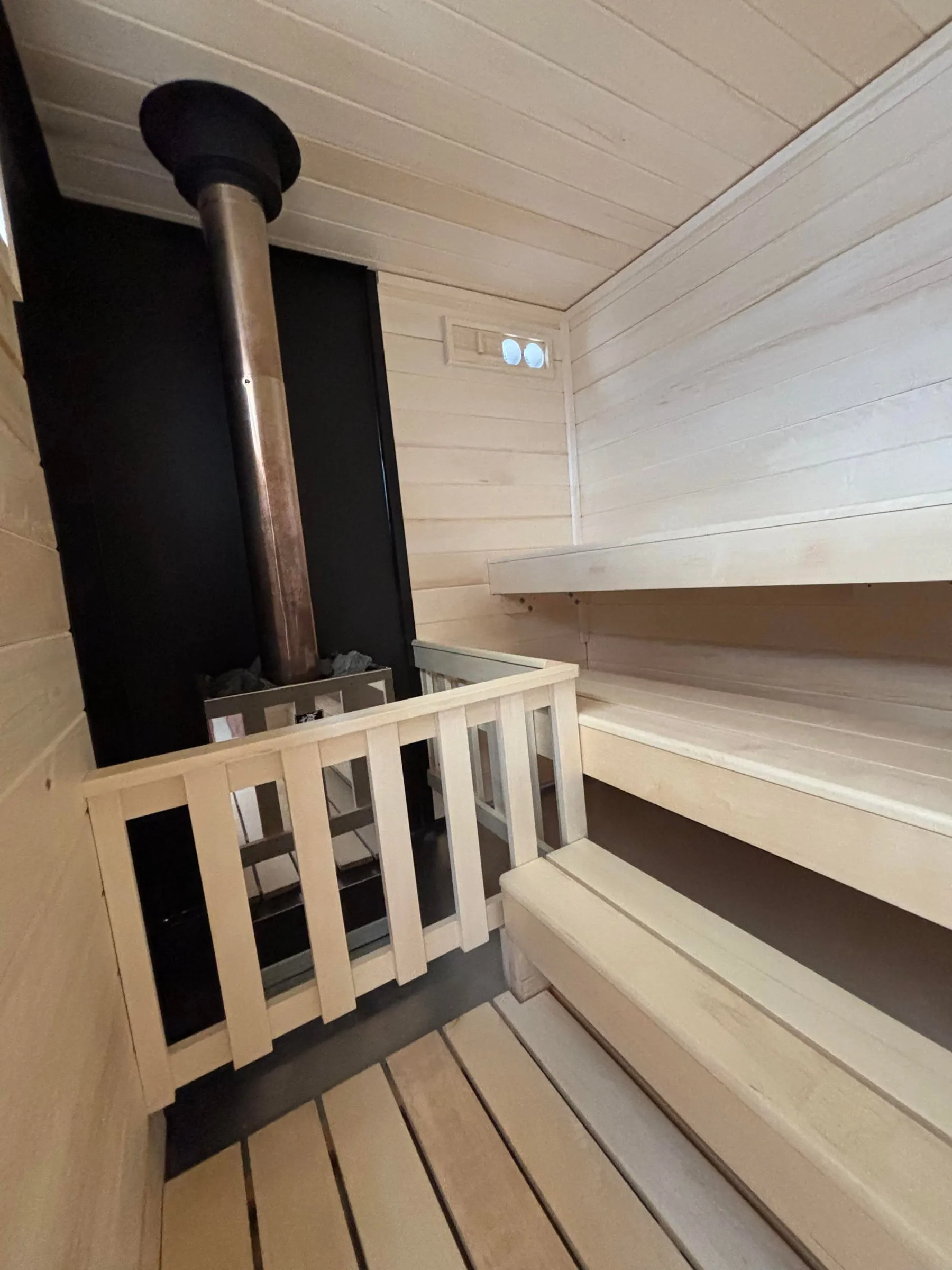 Inside our wood-burning trailer sauna: the chimney pipe rising from the heater to its black ceiling collar, in front of the black non-combustible wall.