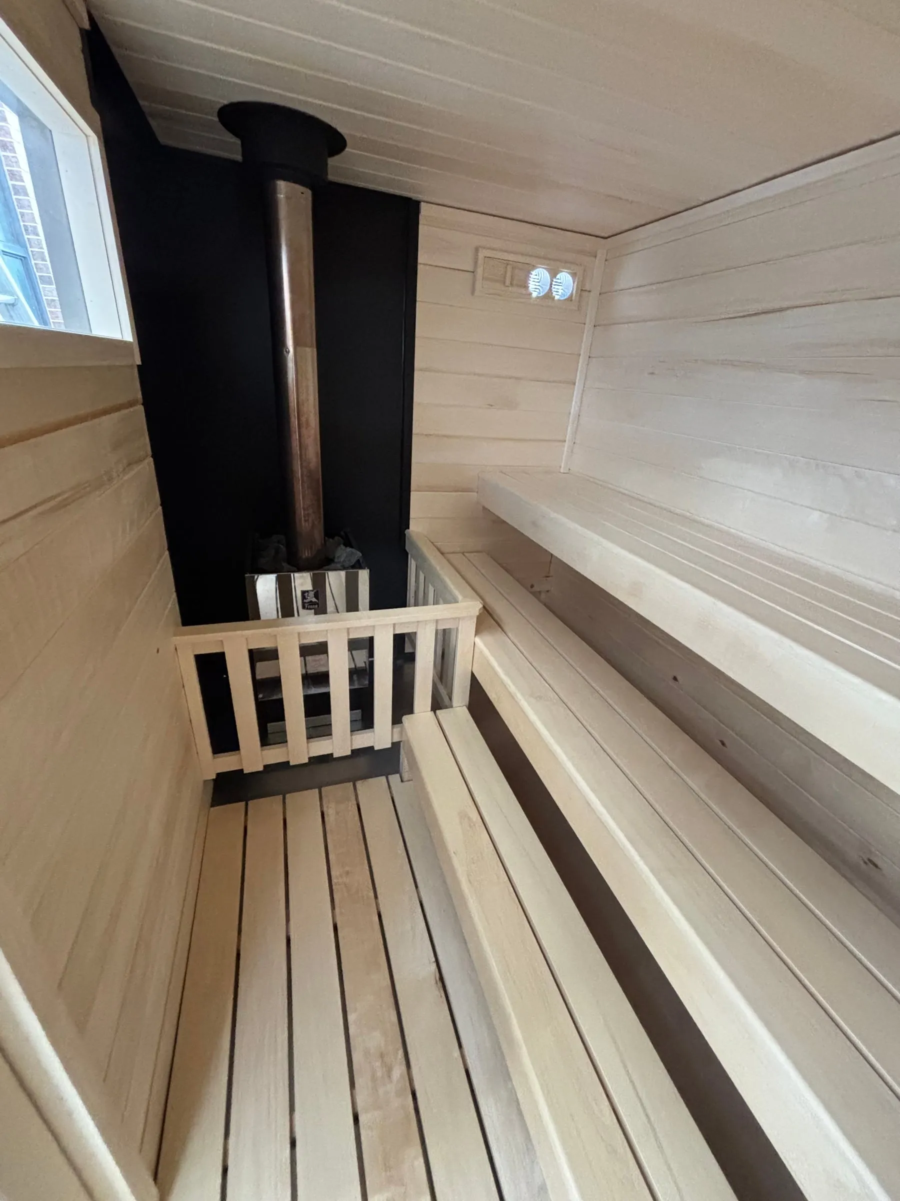 Inside our wood-burning trailer sauna: the stainless steel Fenno wood-burning heater with its sauna rocks, behind an aspen guard rail, with the chimney pipe rising to the ceiling.