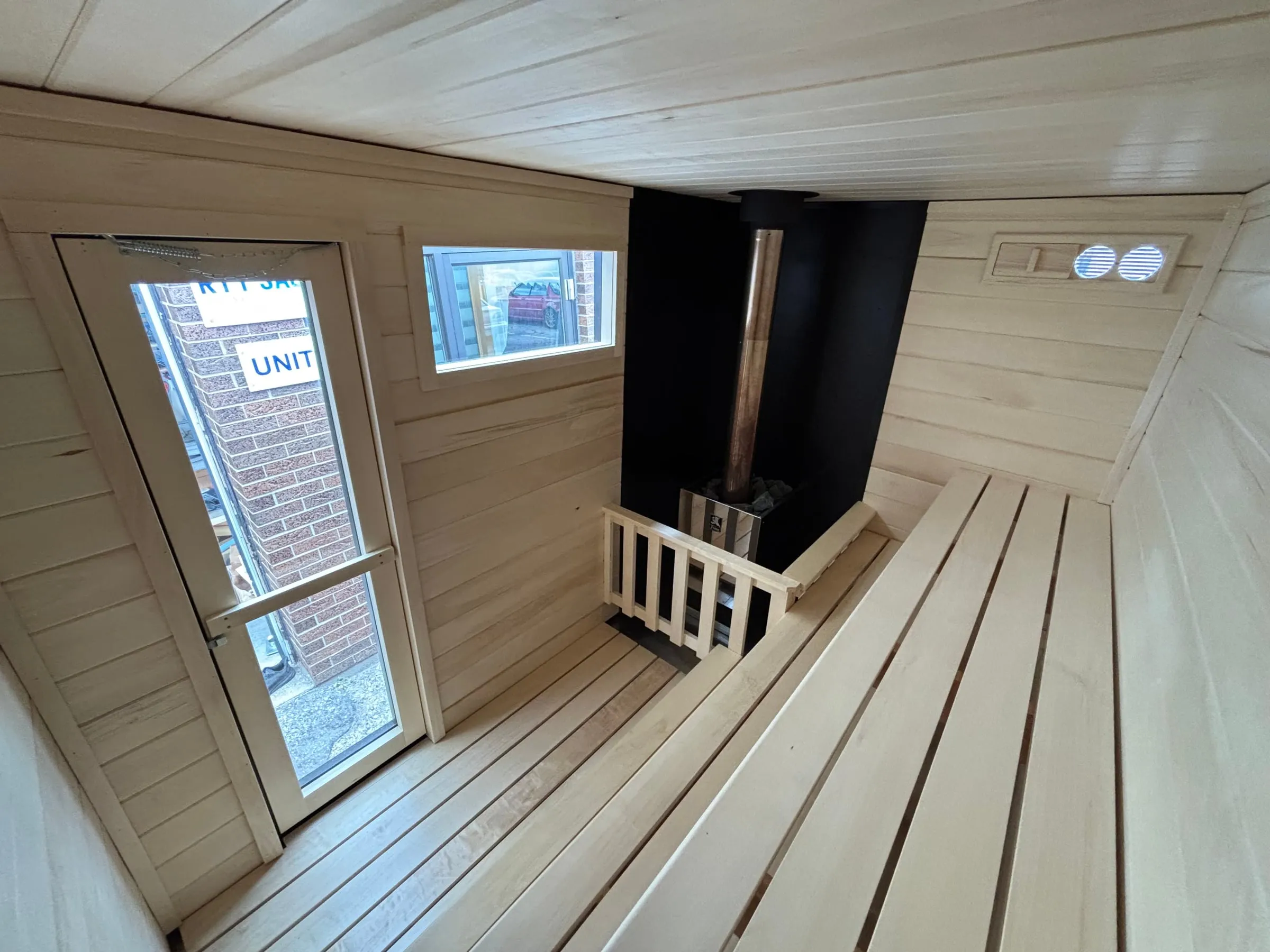 Inside our wood-burning trailer sauna: a full glass door and a window on the end wall, the black non-combustible heater wall with the chimney pipe and vents, and aspen benches.