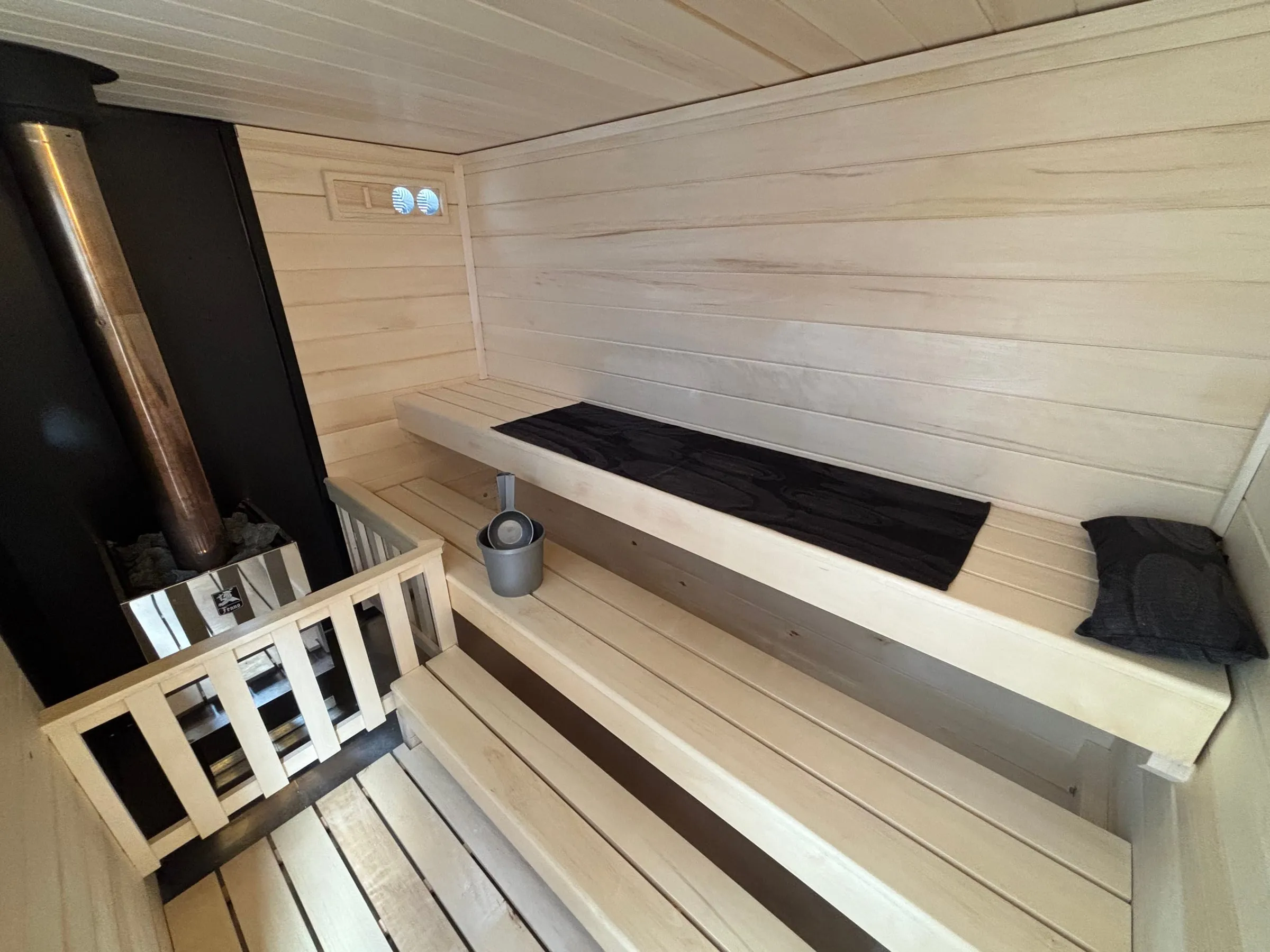Inside our wood-burning trailer sauna: a stainless steel wood-burning heater full of sauna rocks behind an aspen guard rail, a black non-combustible wall and chimney pipe, and two tiers of aspen benches.