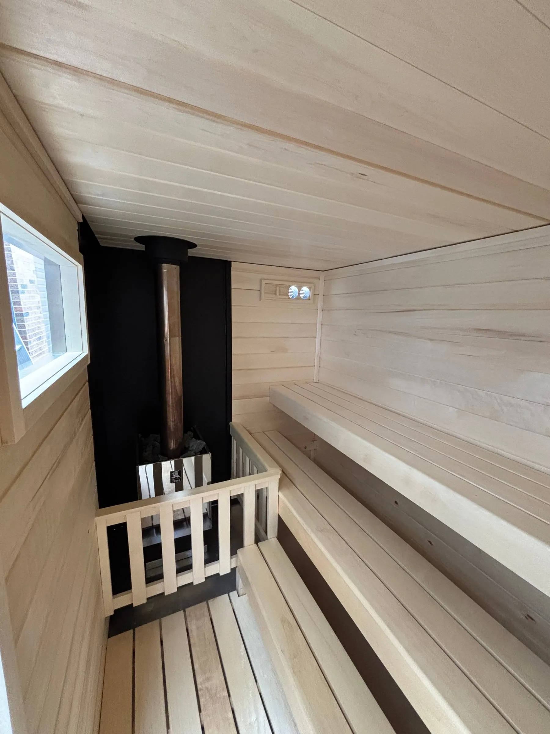 Inside our wood-burning trailer sauna: the chimney pipe rising from the heater to a ceiling collar in front of a black non-combustible wall, with an aspen guard rail and aspen benches.