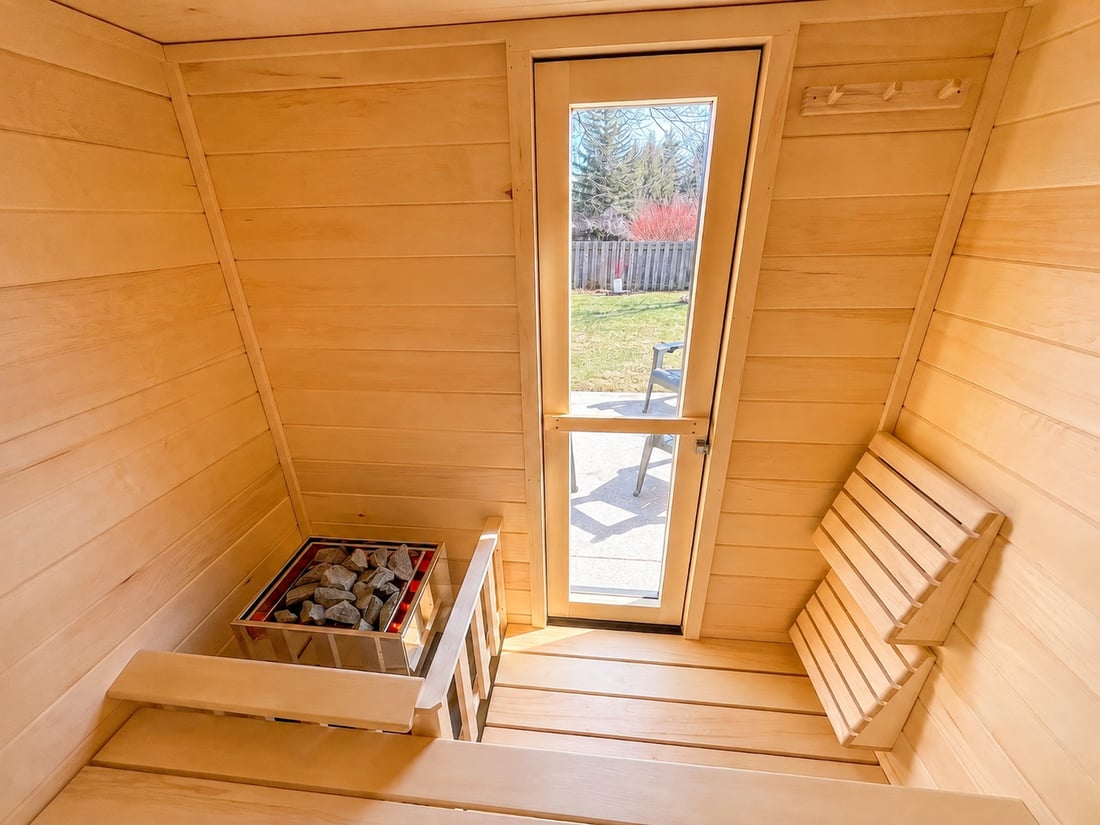 Inside a Ryt Sauna with the heater running: aspen walls, benches and backrest, rocks glowing red in the heater, and the glass door looking out to the yard