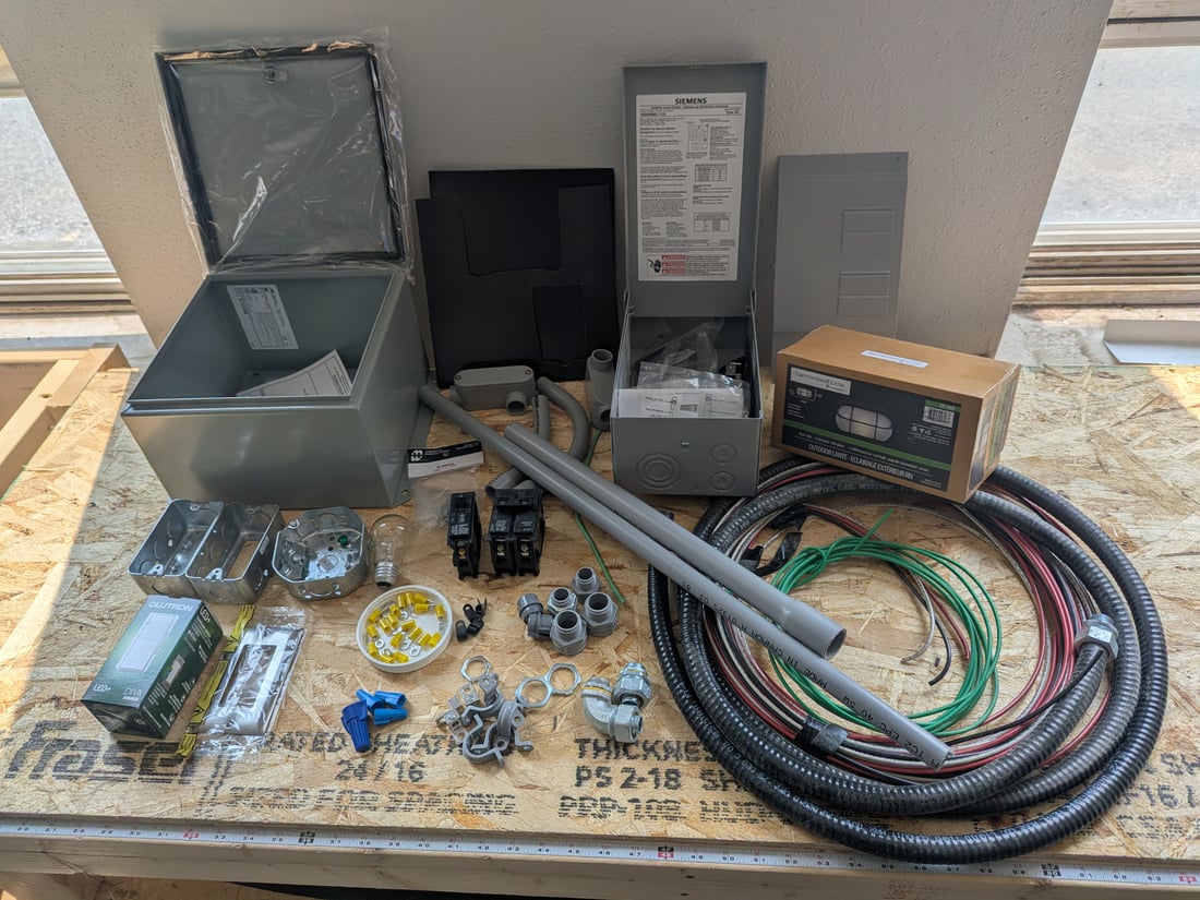 The Option 2 components package laid out on a workbench: a Siemens outdoor sub-panel with its documentation, two grey exterior enclosures, two double-pole breakers, metal device boxes, a Lutron dimmer and cover plate, a boxed outdoor light fixture, conduit lengths and fittings, wire connectors, and coiled red, black, green and white wire with flexible metal conduit