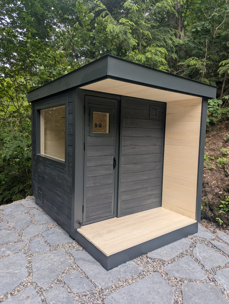 A Ryt Sauna in dark Chamclad cladding with a black aluminum roof, an aspen-lined covered entry with aspen deck boards, a glass-panelled door set under the overhang, and a wide horizontal window on the adjoining wall